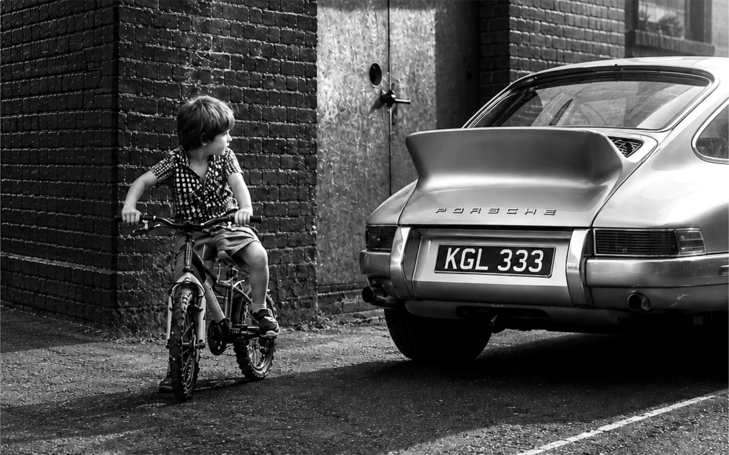 A boy on a bicycle looking back at a classic Porsche parked in a brick alley